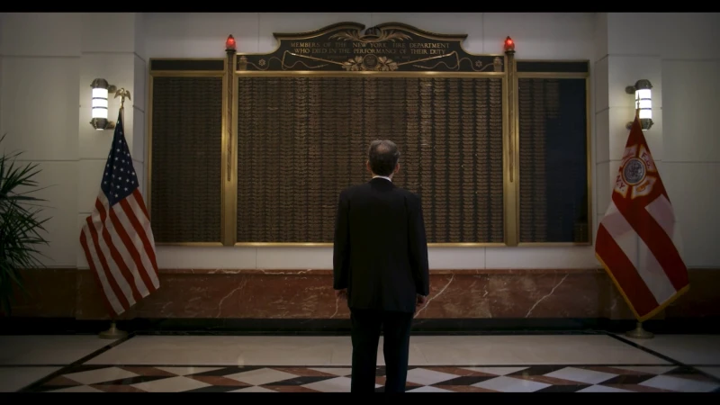 A man in a dark suit stands facing a large memorial plaque flanked by flags and wall sconces inside a grand interior space.