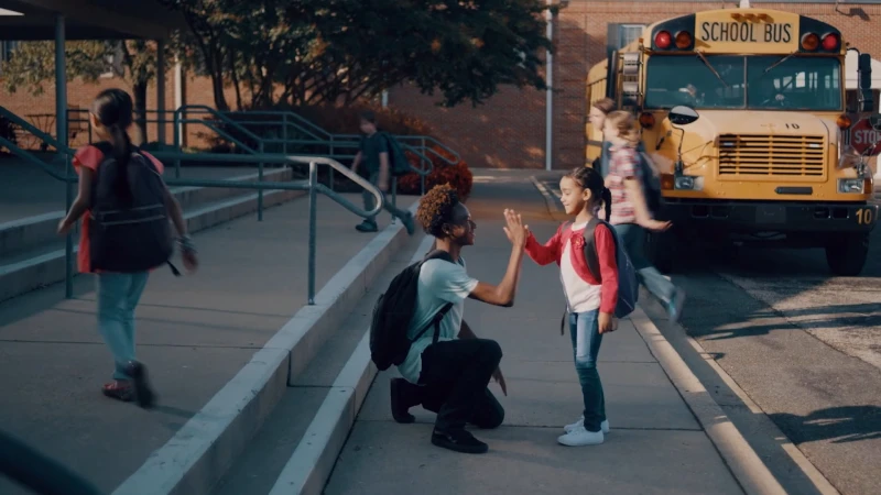 Young Black man kneeling on pavement high-fiving a young girl with a yellow school bus parked in the background.