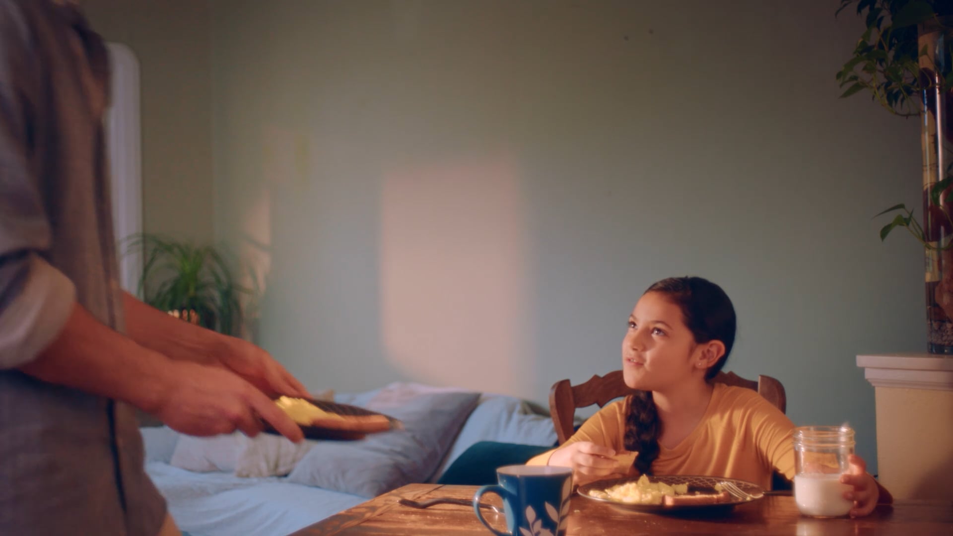 Young girl seated at a wooden table looking up as a person serves food on a plate in a domestic bedroom interior.