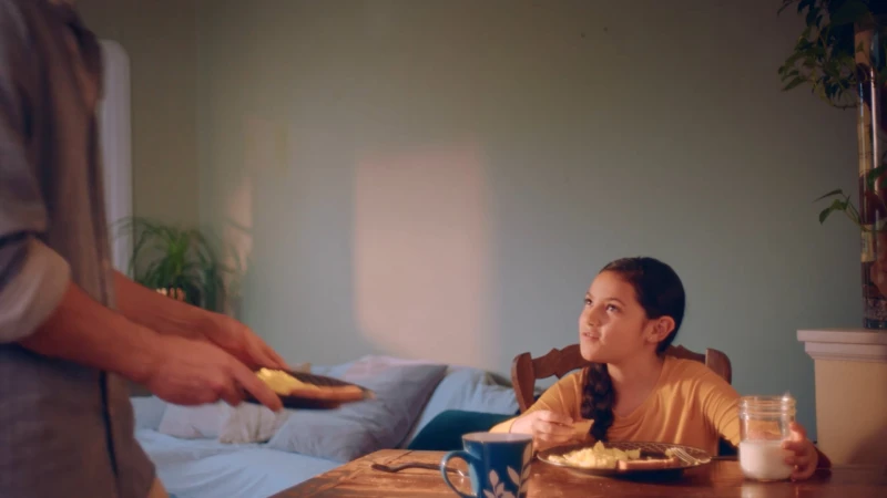 Young girl seated at a wooden table looking up as a person serves food on a plate in a domestic bedroom interior.