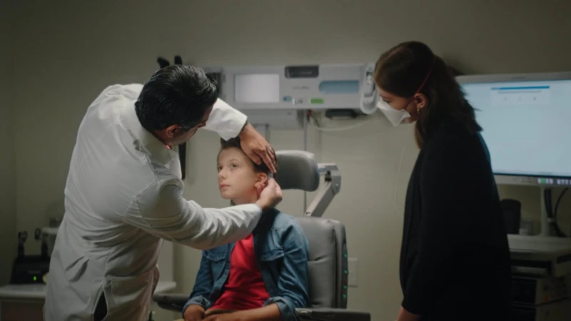 Doctor examining a young boy's ear in a medical clinic while a woman stands nearby