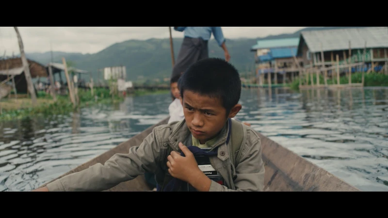 Young boy in a jacket seated in a wooden boat on a river with stilt houses on the water and mountains in the background