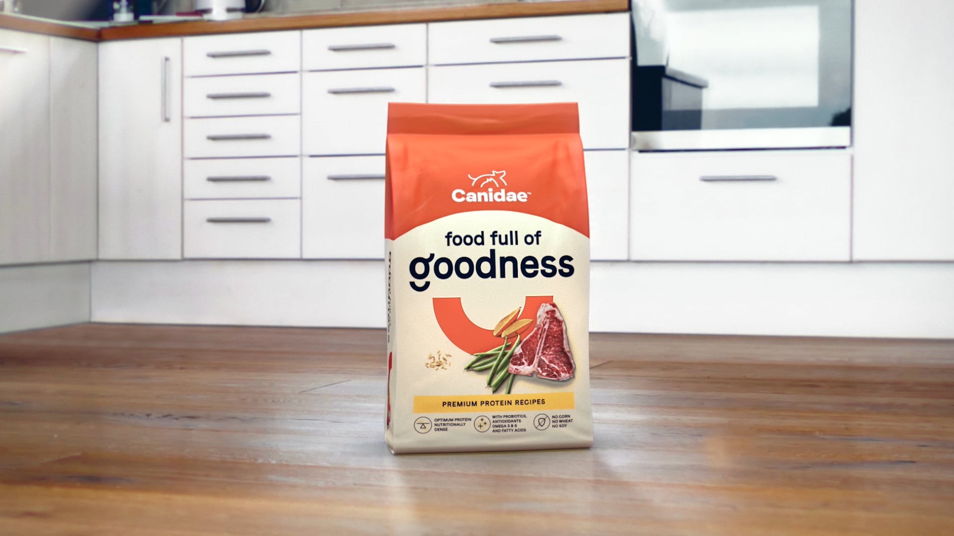 A bag of Canidae dog food stands upright on a wooden floor in a modern white kitchen.