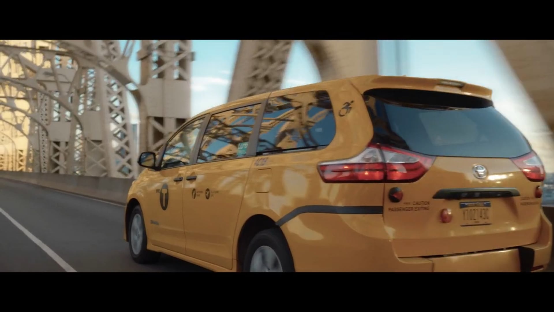 Yellow taxi minivan driving across a bridge with steel truss architecture visible in the background.