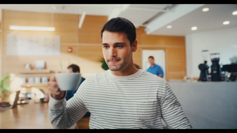 Man in a striped sweater holding a white coffee cup in a cafe setting filmed by Erin Collett