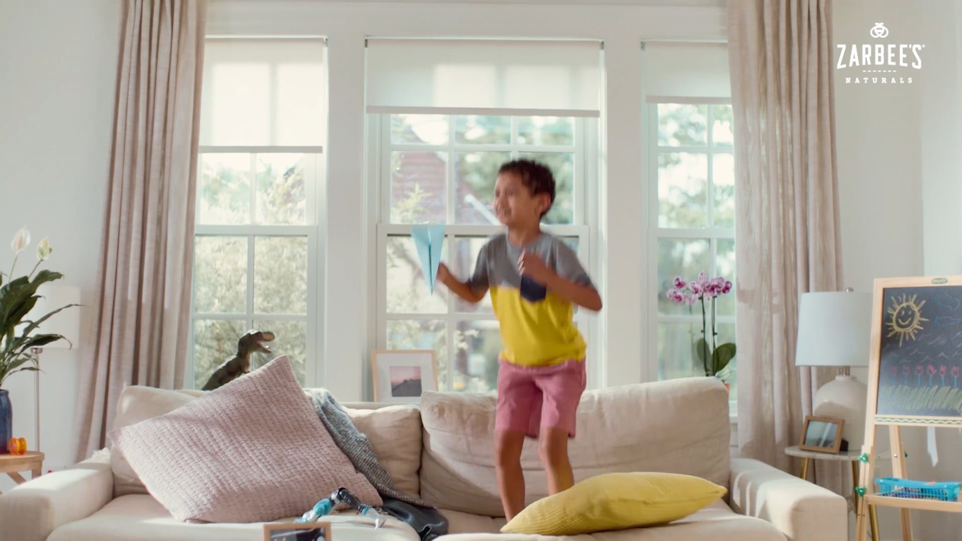 A young boy jumping on a cream sofa in a living room, holding a blue paper airplane.