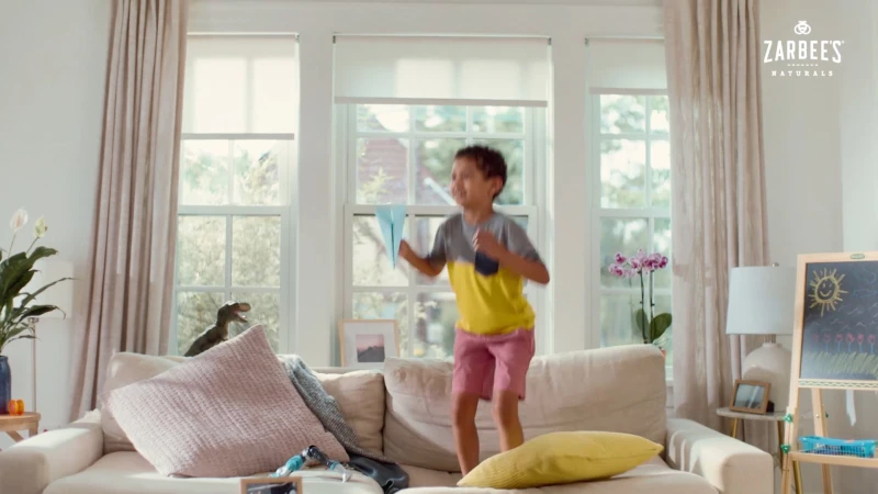 A young boy jumping on a cream sofa in a living room, holding a blue paper airplane.