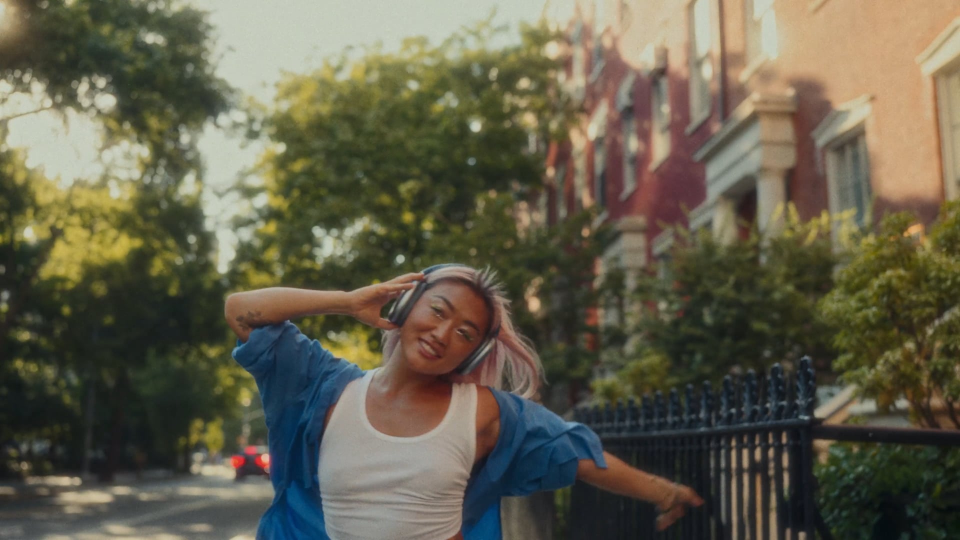 A smiling young woman with pink-streaked hair wearing a blue shirt and white tank top listens to over-ear headphones on a city street.