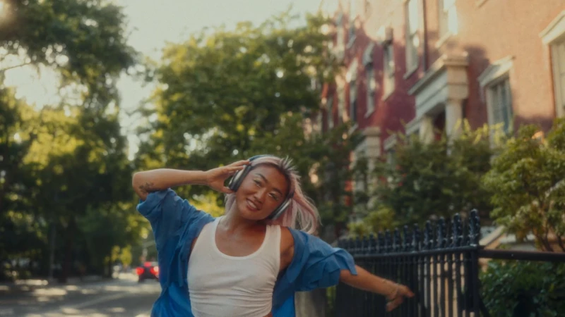 A smiling young woman with pink-streaked hair wearing a blue shirt and white tank top listens to over-ear headphones on a city street.