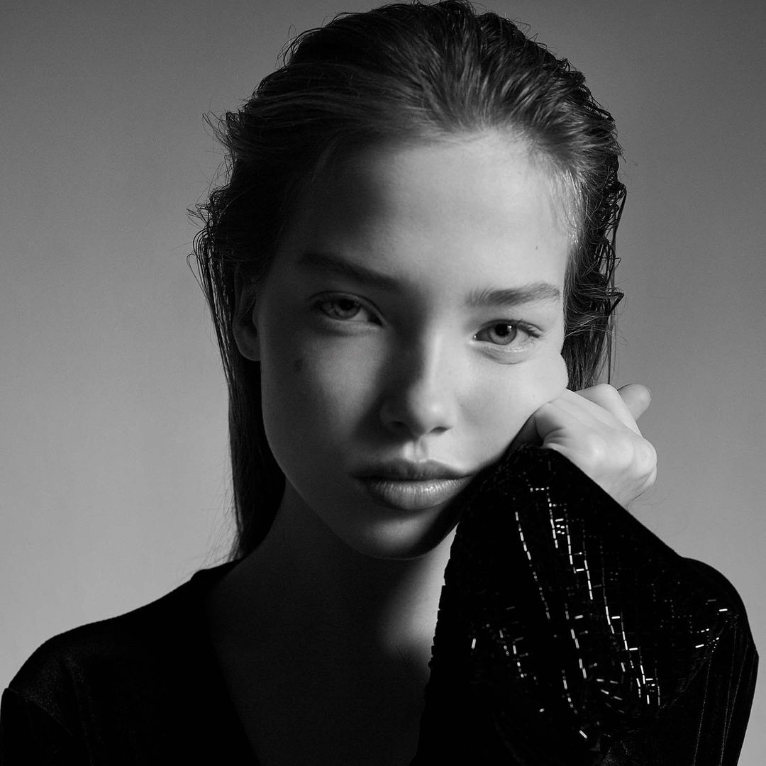 Monochrome portrait of a young woman with slicked-back hair and hand to face, wearing a dark shimmering top against a grey background.