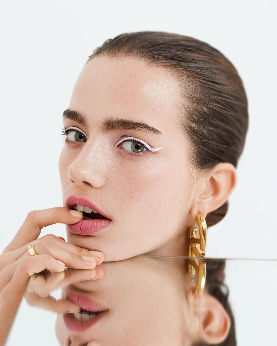 Close-up beauty shot of an adult woman with white eyeliner, gold hoop earrings, and her finger on her lip, reflected in glass.