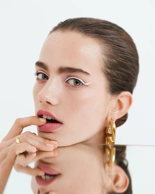 Close-up beauty shot of an adult woman with white eyeliner, gold hoop earrings, and her finger on her lip, reflected in glass.