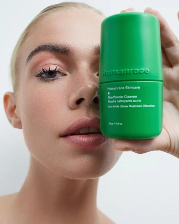 Close-up of a fair-skinned woman holding a bright green Humanrace skincare product in front of her face in a studio setting.