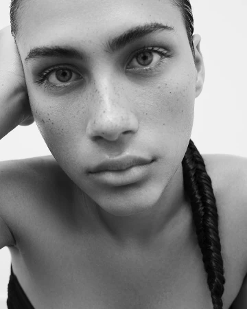 Close-up portrait of a woman with freckles and a dark braided ponytail looking directly at the camera by Paula Latimori.