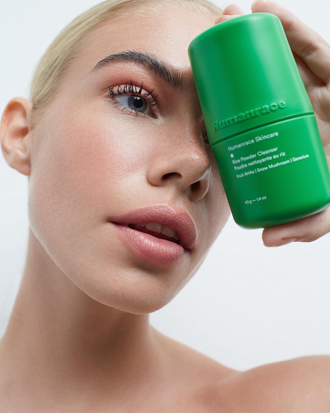 Close-up of a fair-skinned woman holding a green Humanrace skincare product against a neutral background, promoting beauty and skincare.