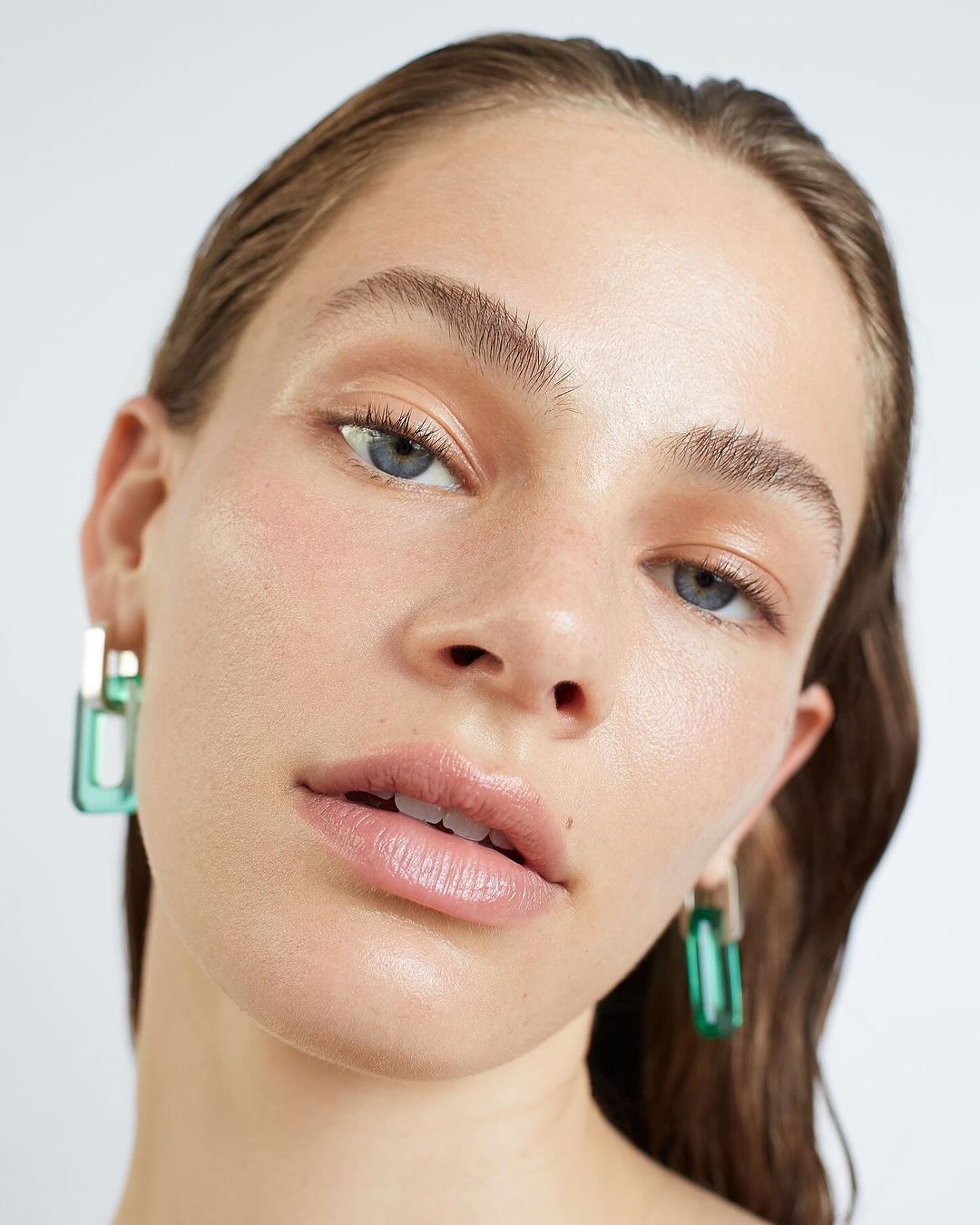 Close-up studio shot of an adult woman with fair skin wearing green earrings, with a neutral expression and minimal makeup.