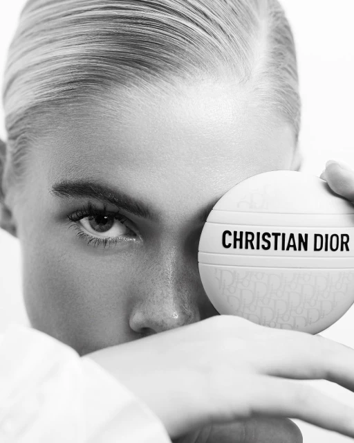 Black-and-white beauty shot of a woman holding Christian Dior makeup, showing her eye, eyebrow, and part of her face and shoulder.