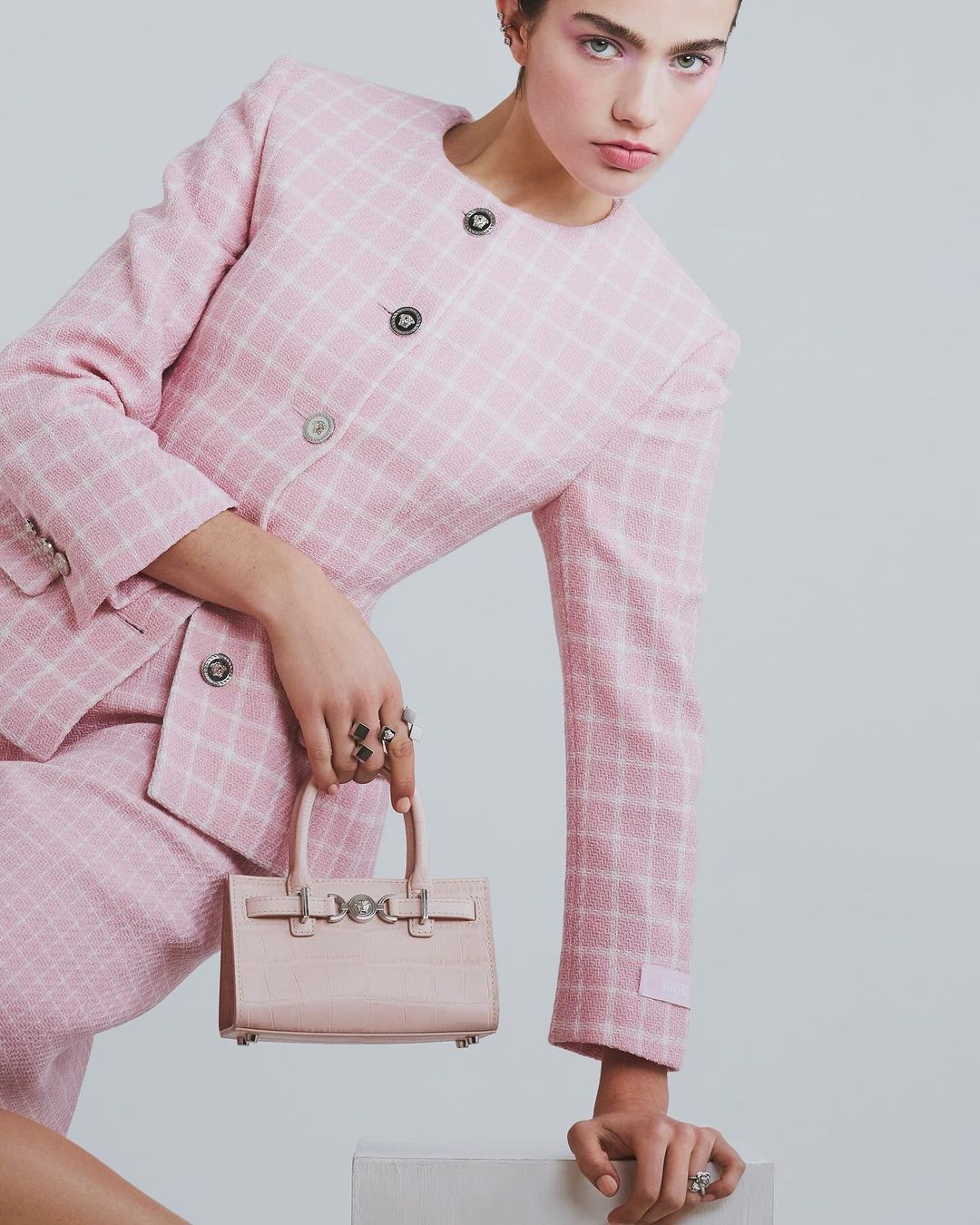 A young woman poses in a pink tweed skirt suit with a matching mini handbag against a neutral background, looking at the camera.