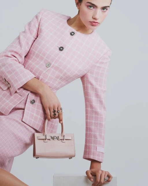 A young woman poses in a pink tweed skirt suit with a matching mini handbag against a neutral background, looking at the camera.