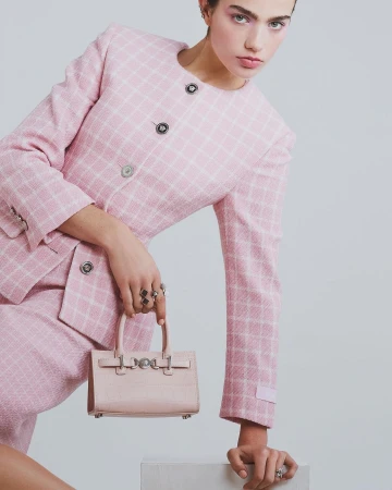A young woman poses in a pink tweed skirt suit with a matching mini handbag against a neutral background, looking at the camera.