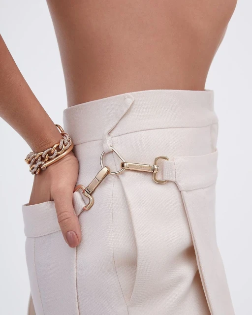 Close-up shot of a female wearing cream-colored pants with metallic hardware, showing a bracelet on her wrist against a light background.