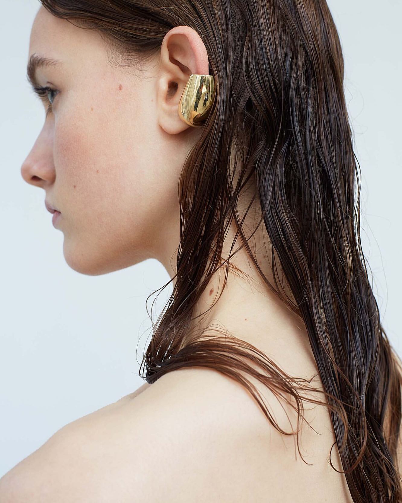 Close-up studio shot of a young adult female model wearing a large, modern gold ear cuff earring and wet, dark brown hair against a white backdrop.