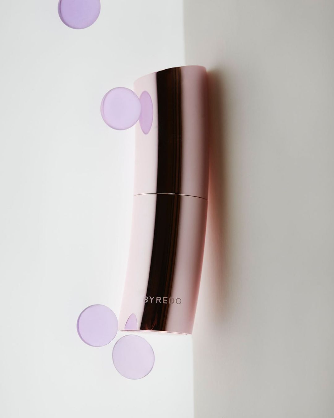 Pink curved perfume bottle by Byredo against a white background with floating translucent purple circles.
