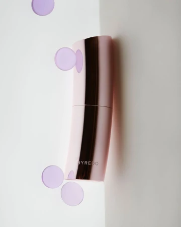 Pink curved perfume bottle by Byredo against a white background with floating translucent purple circles.