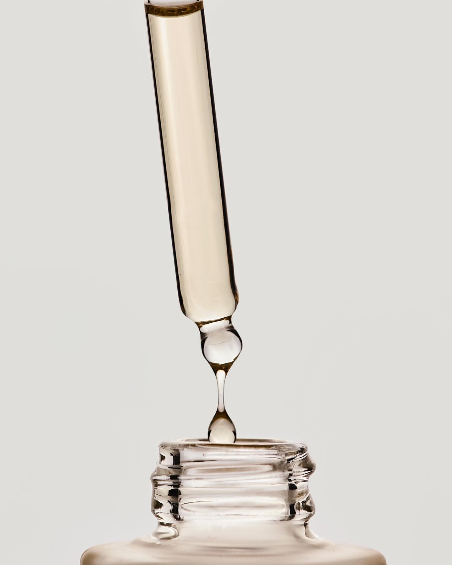 Close-up studio shot of a glass dropper dispensing cosmetic serum into a bottle against a plain background, showcasing skincare product.