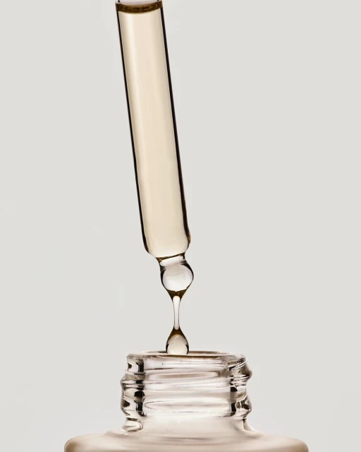 Close-up studio shot of a glass dropper dispensing cosmetic serum into a bottle against a plain background, showcasing skincare product.