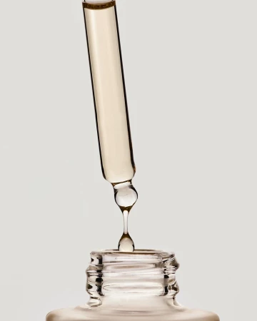 Close-up studio shot of a glass dropper dispensing cosmetic serum into a bottle against a plain background, showcasing skincare product.