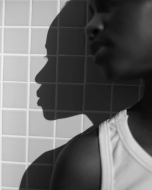 Monochrome portrait of an adult with short dark hair wearing a white tank top, side lit against a tiled background, featuring their shadow.