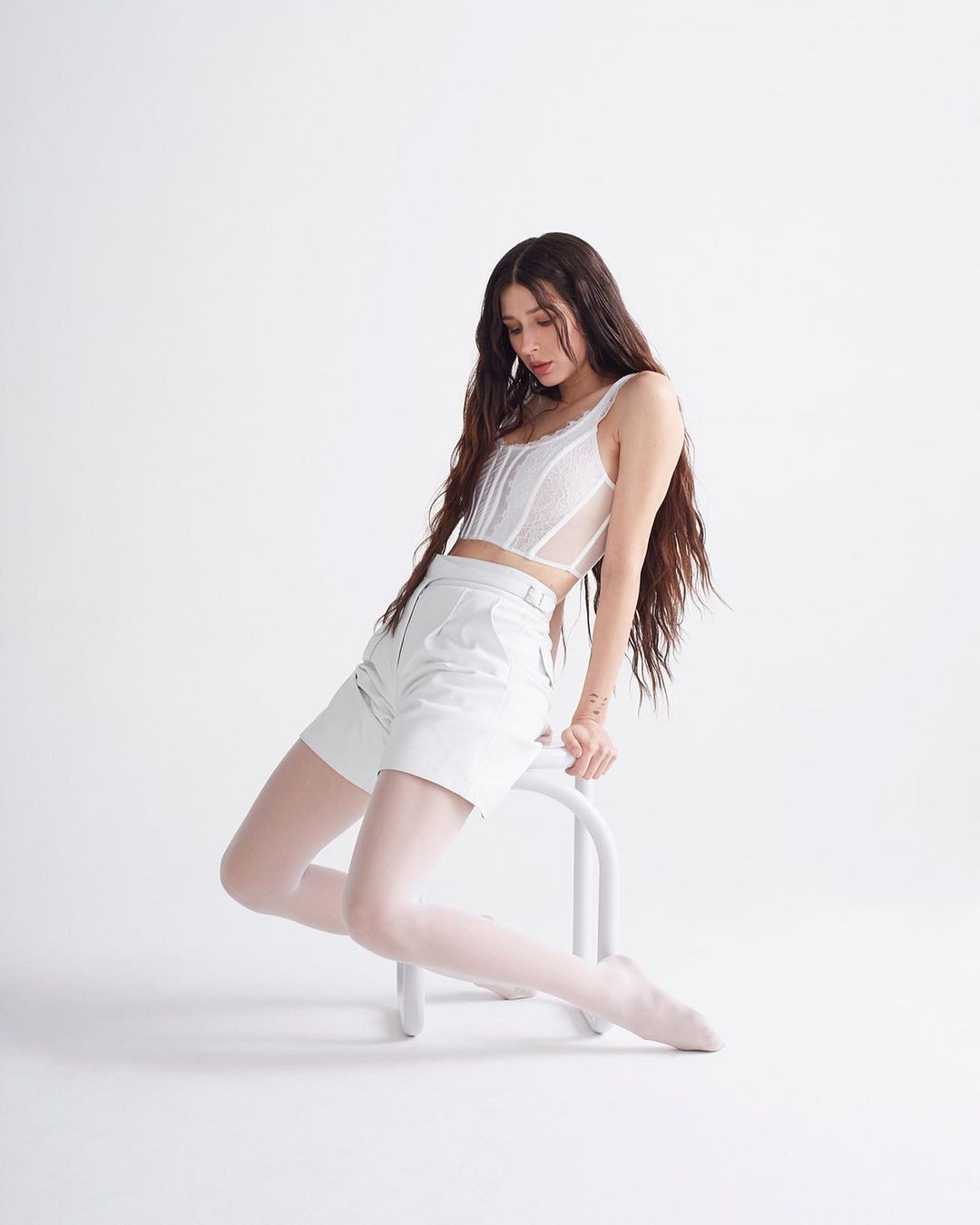 A young brunette woman with long hair poses indoors on a white stool wearing white shorts, sheer tights, and a white tank top.
