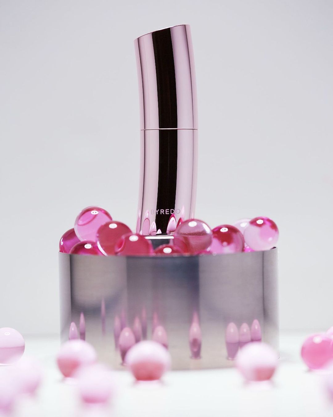 A pink Byredo lipstick tube is displayed on a reflective chrome platform surrounded by translucent pink beads against a white background.