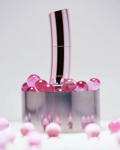 A pink Byredo lipstick tube is displayed on a reflective chrome platform surrounded by translucent pink beads against a white background.