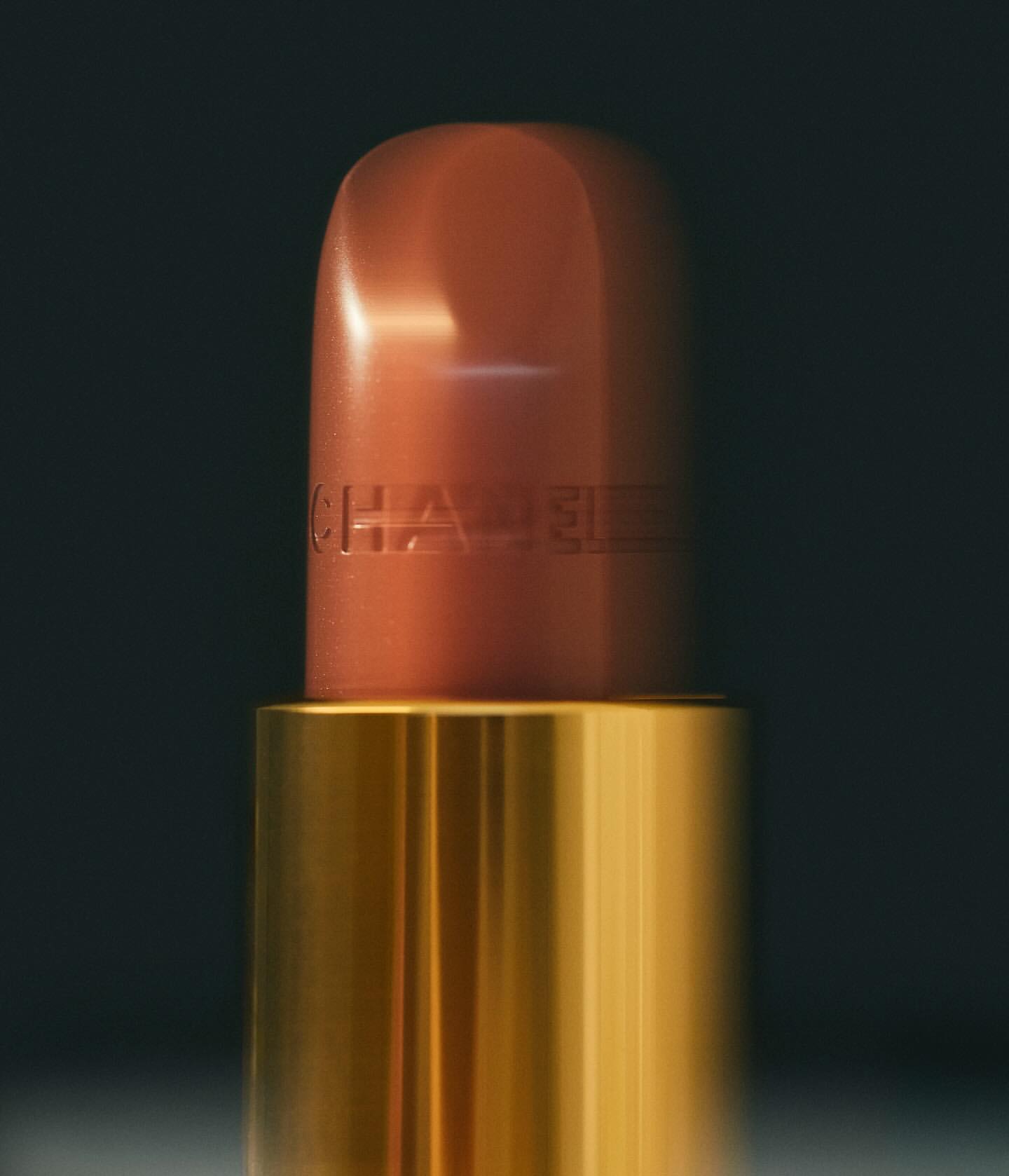 Close-up of a light-brown Chanel lipstick with a gold case against a dark background, highlighting the brand and makeup product.