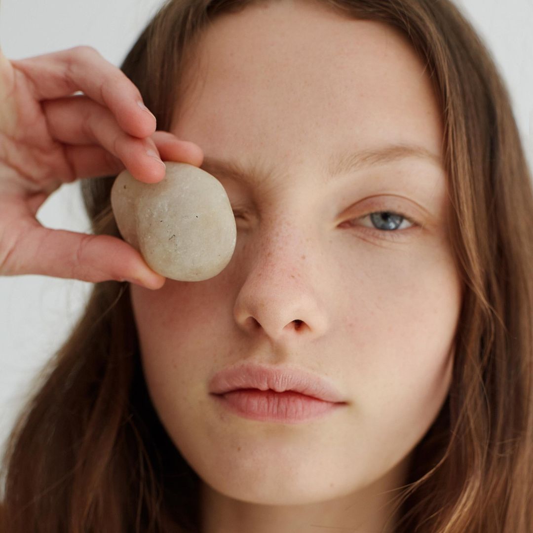 Close-up of a young woman holding a smooth stone over her eye, showcasing a minimalist beauty concept in a bright, clean studio environment.