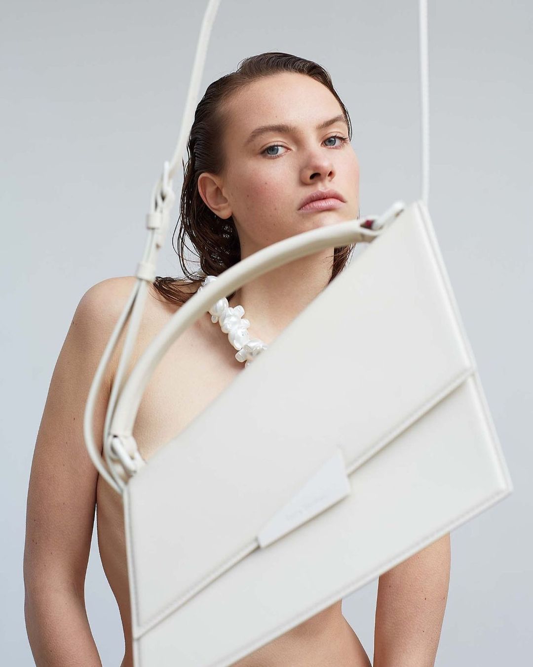 Woman in fashion shot with a white sculptural necklace and a white handbag against a white background in a studio.