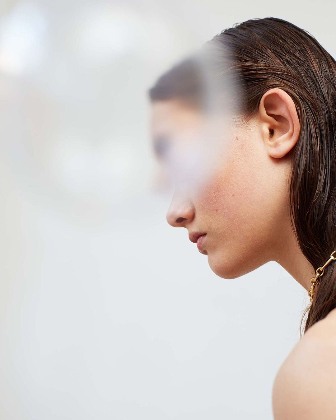 Close-up profile of an adult woman with wet, dark hair, wearing a gold chain necklace against a bright white, blurred background.