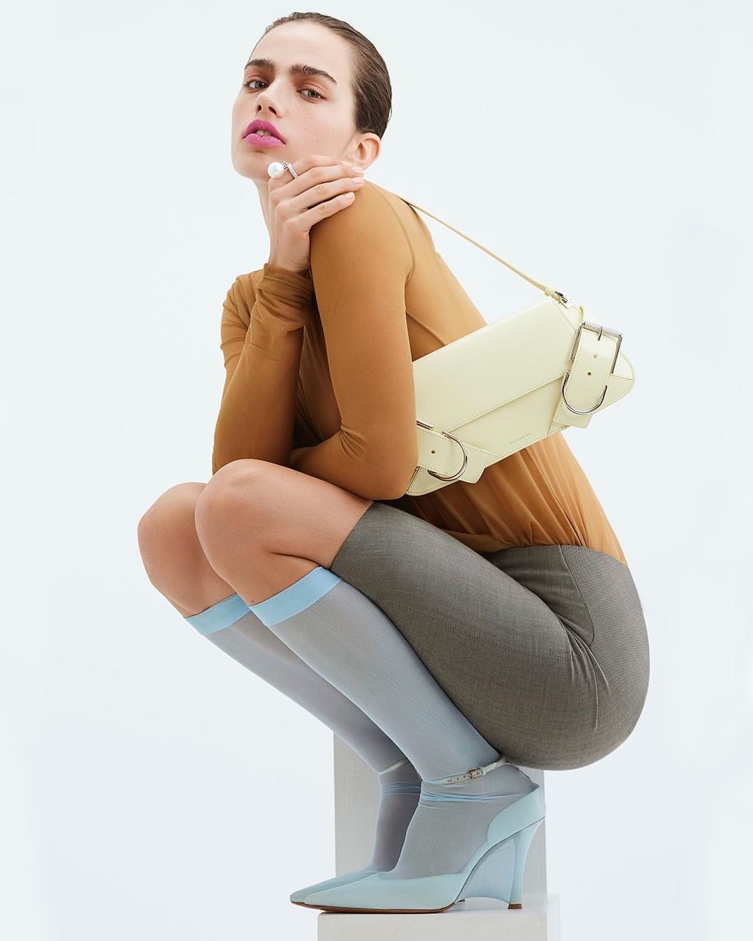 Fashion shot of a woman in hosiery and wedge heels posing with a shoulder bag against a white backdrop in a photo studio.