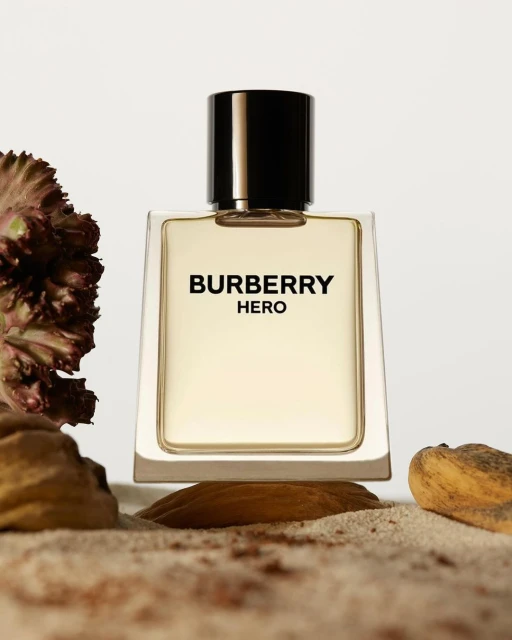Burberry Hero perfume bottle on sandy surface with botanical elements against a white backdrop in a studio product shot.