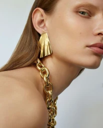 Close-up portrait of a woman wearing large gold earrings and a heavy gold chain necklace against a light background.
