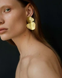 Close-up portrait of a woman wearing large sculptural gold earrings against a dark background, photographed by Paula Latimori.