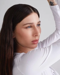 Close-up portrait of a woman with long brown hair wearing a white top against a clean light background, photographed by Paula Latimori