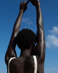 Rear view of a Black woman with short dark hair raising her arms against a clear blue sky, wearing a white top.