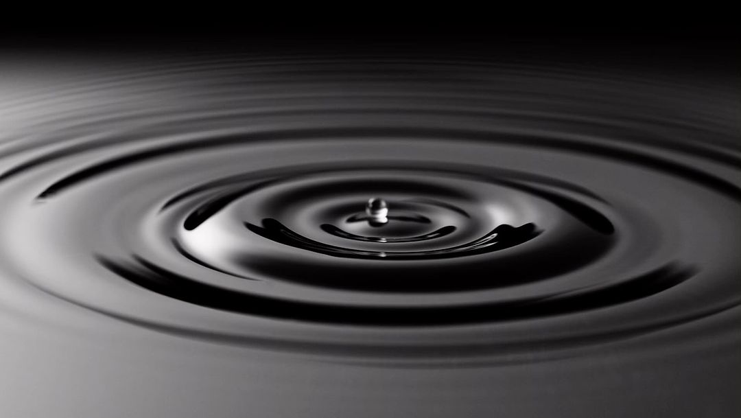 Water drop creates concentric ripples on the liquid surface in a dark setting, reflecting the impact and circular wave patterns.