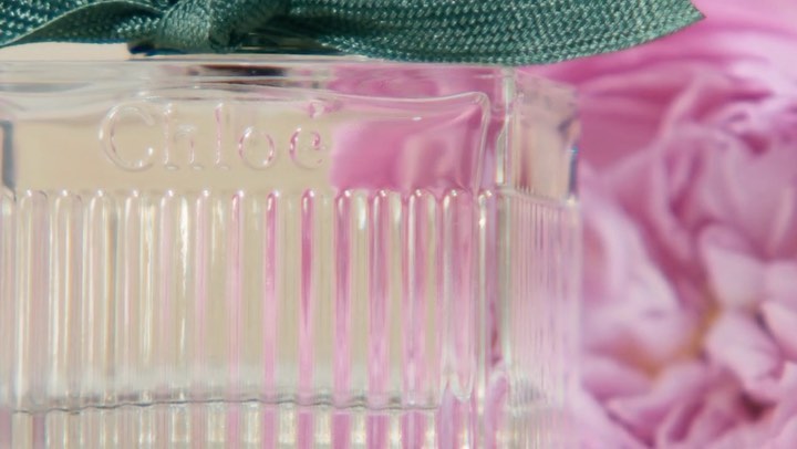 Close-up of a Chloe perfume bottle with a green ribbon, set against a blurred pink floral background. Product photography.