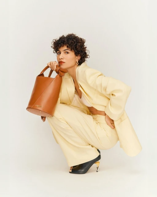 A woman with curly hair poses crouching in a yellow suit with a tan bucket bag in a bright, eye-level studio shot.
