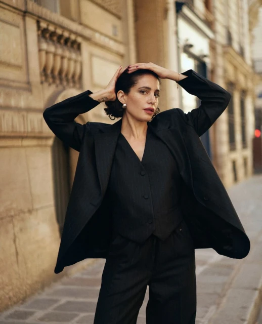 Adult female poses outdoors in dark business suit, vest, and trousers, hands behind head, on city street with tall beige buildings.