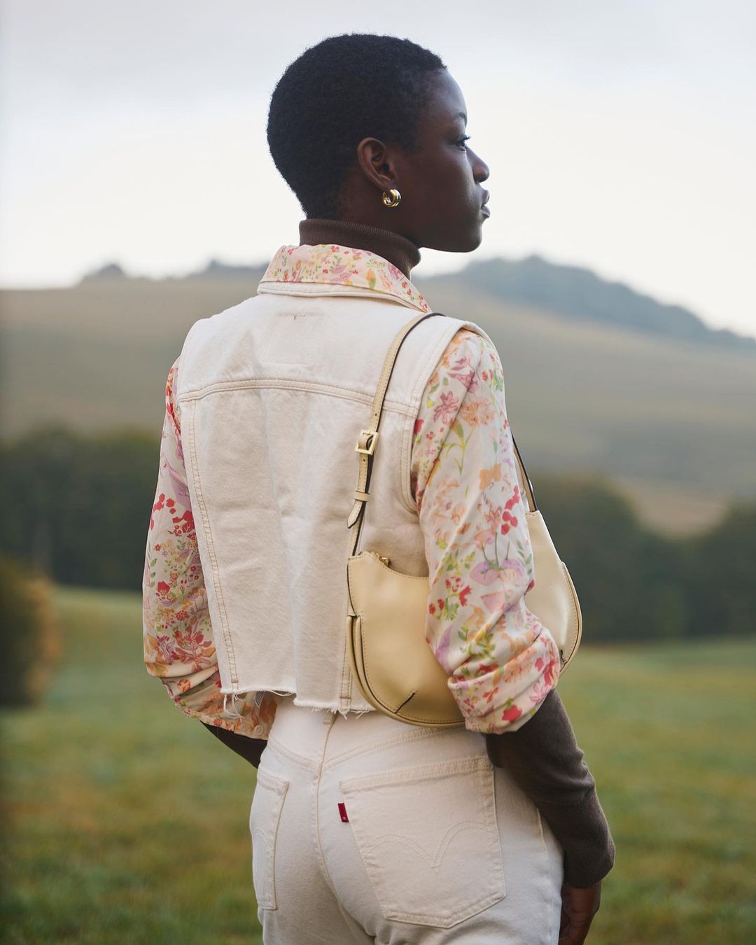 Woman in casual fashion stands in an outdoor setting with rolling hills; she wears light-wash jeans and vest, a floral top, and a purse.